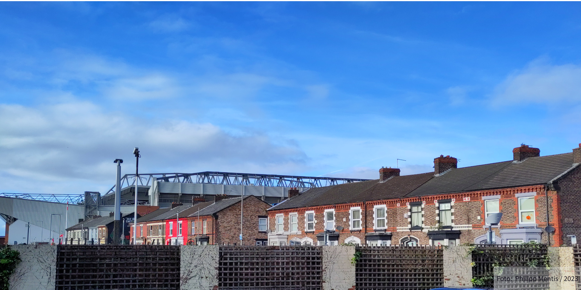 !B Behind a common Liverpool street lined with houses, the Anfield ground is visible.